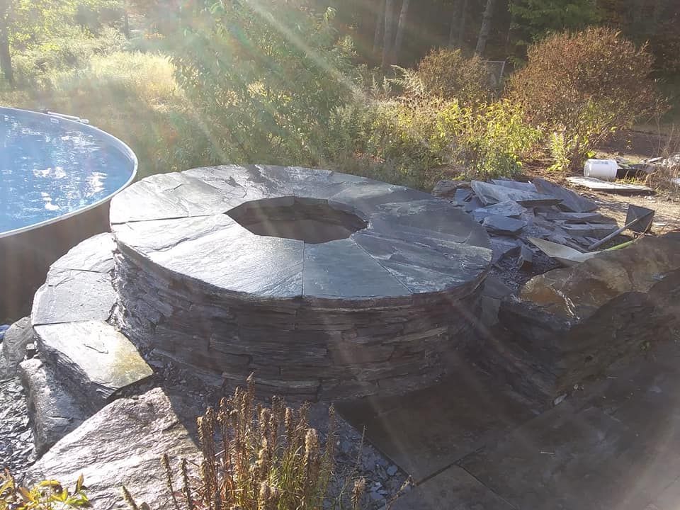 Stone fire pit with built-in seating, near a metal pool, in a natural outdoor setting.