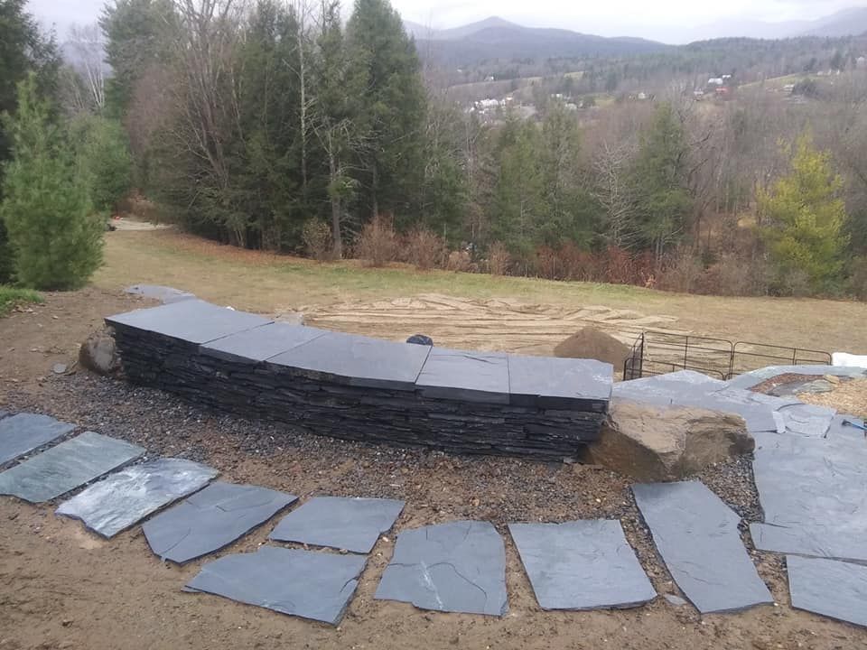 Stacked dark slate pavers with scattered stones on a dirt path, leading to a grassy hill and distant trees.