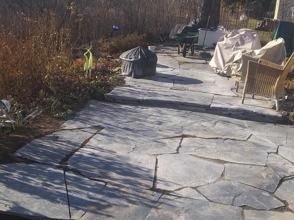 Stone patio with cracks, leading to a yard with bushes, covered furniture, and a fire pit.