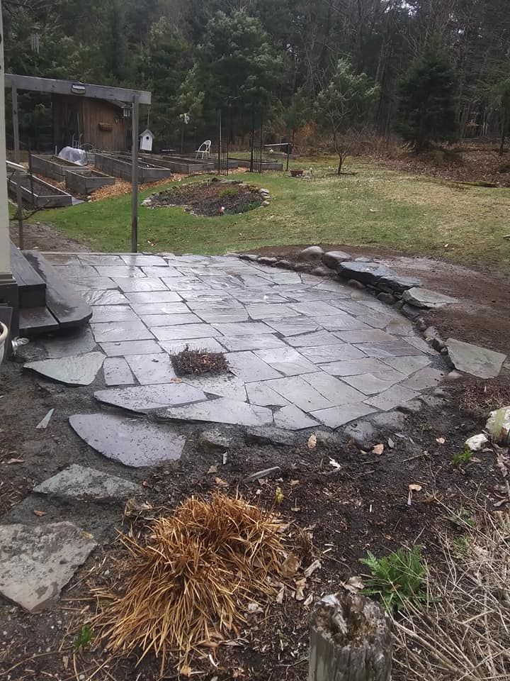 Stone patio with irregular-shaped pavers and border, set in a muddy yard, near grass and trees.