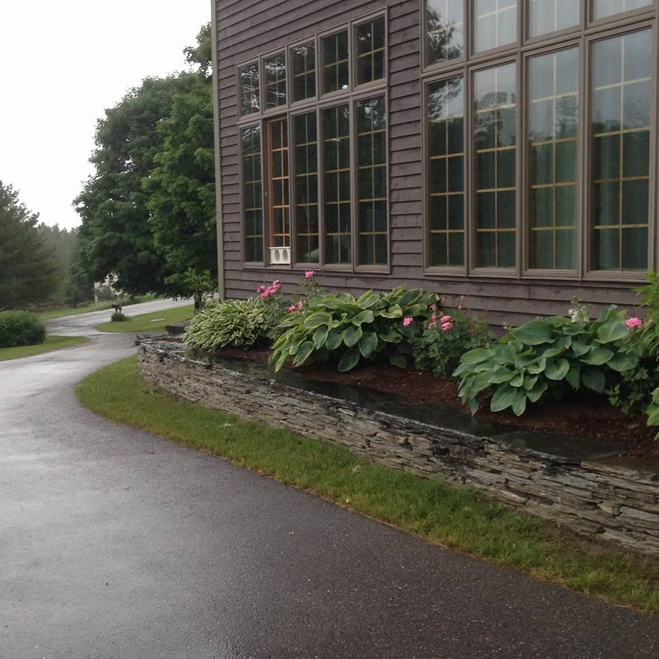 Exterior of a building with tall windows, next to a stone wall and a paved pathway. Lush green plants.