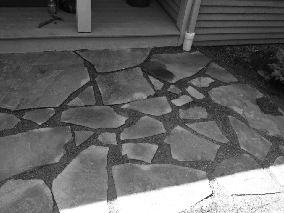 Black and white photo of a flagstone patio leading to a doorway, with dark gravel in between the stones.