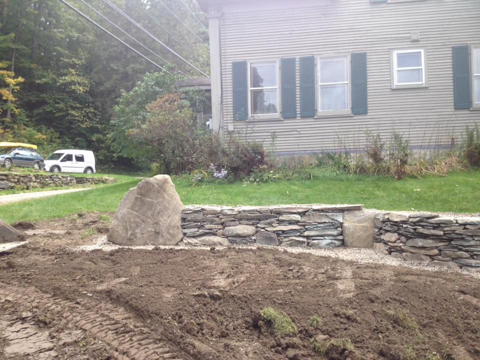 A large rock and low stone wall in front of a house, with dirt and grass, and vehicles in the distance.