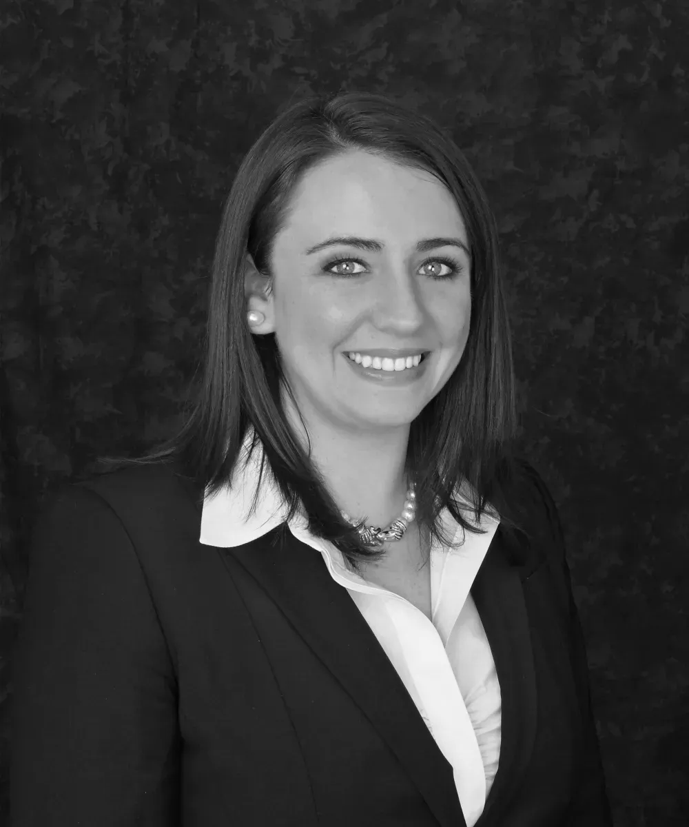 Woman in a black suit smiles at the camera; dark background.