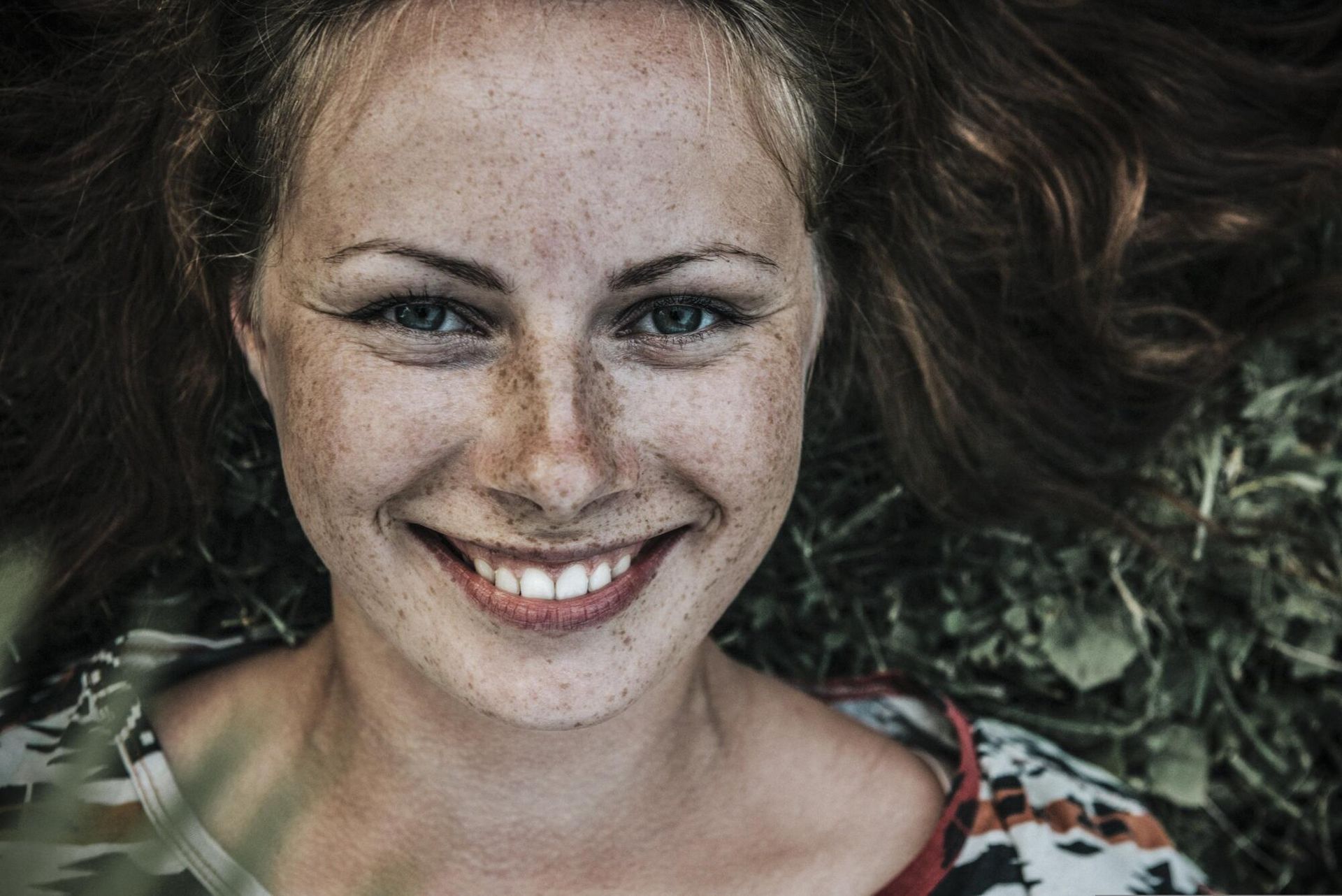 A woman with freckles is smiling while laying in the grass.