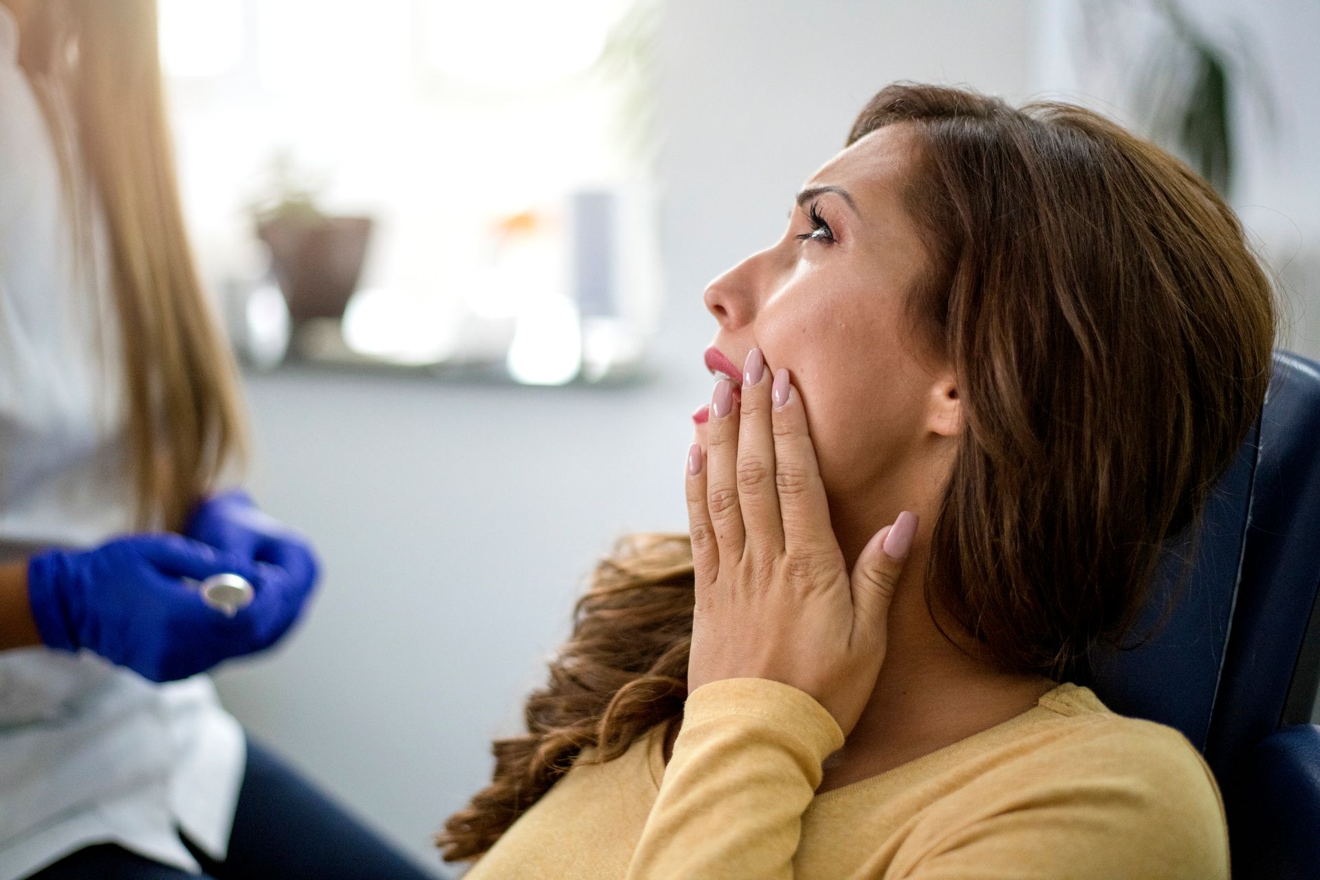 Woman in dental chair, hand on cheek, showing pain. Dentist in blue gloves, holding tools.