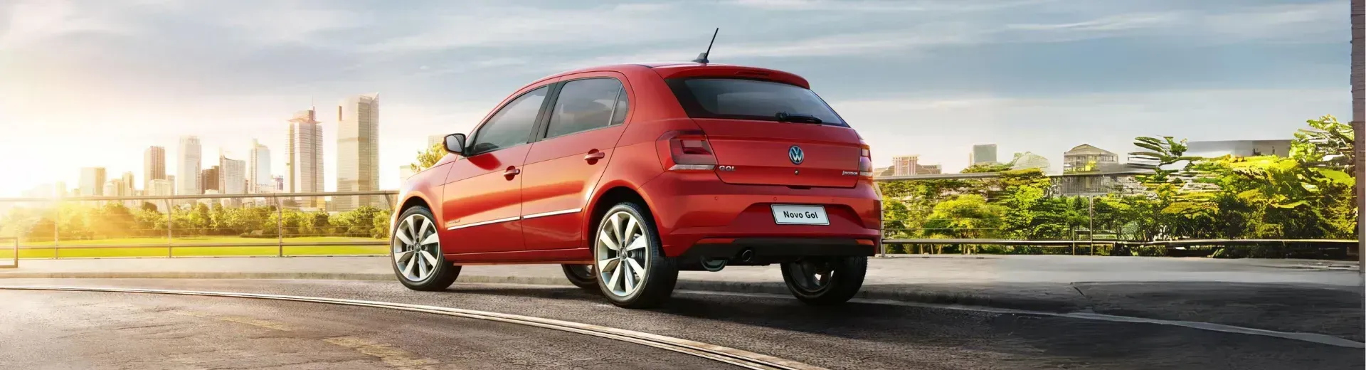 Carro hatchback vermelho dirigindo em uma estrada com o horizonte da cidade ao fundo em um dia ensolarado.