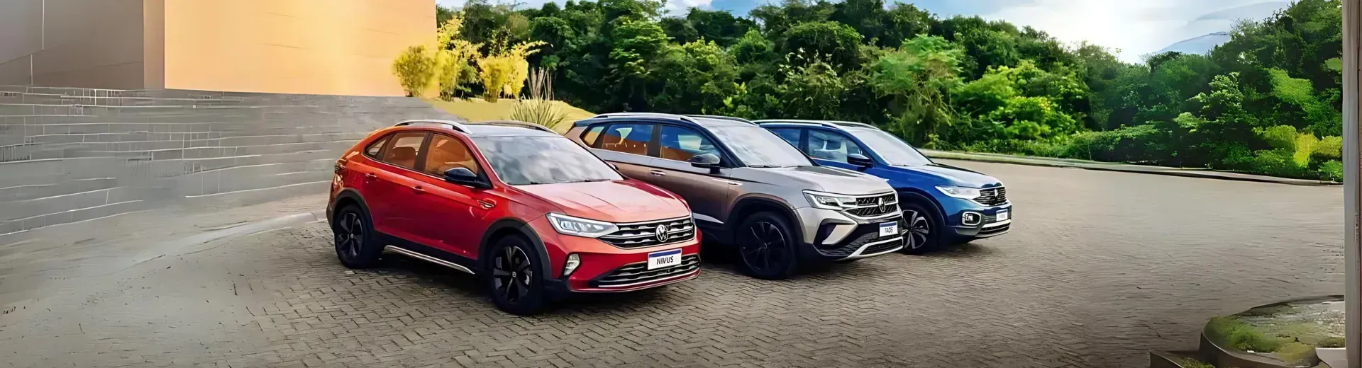 Três SUVs, uma vermelha, uma cinza e uma azul, estacionadas em um terreno pavimentado com vegetação exuberante ao fundo.