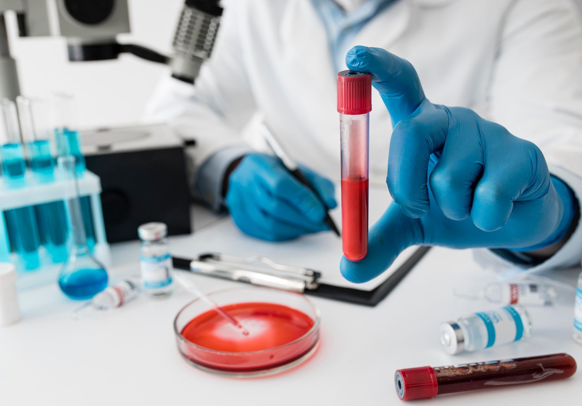 Scientist in lab coat and gloves holds a blood sample tube. Other lab equipment, including a microscope, on the table.