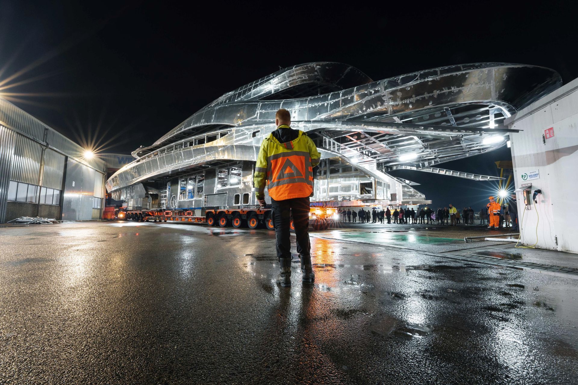 Een man in een oranje jasje staat 's nachts voor een groot gebouw.
