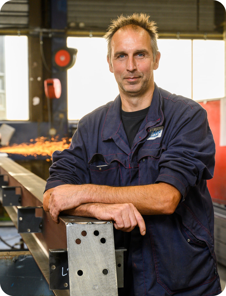 Een man in een blauw uniform staat met zijn armen over elkaar in een fabriek.