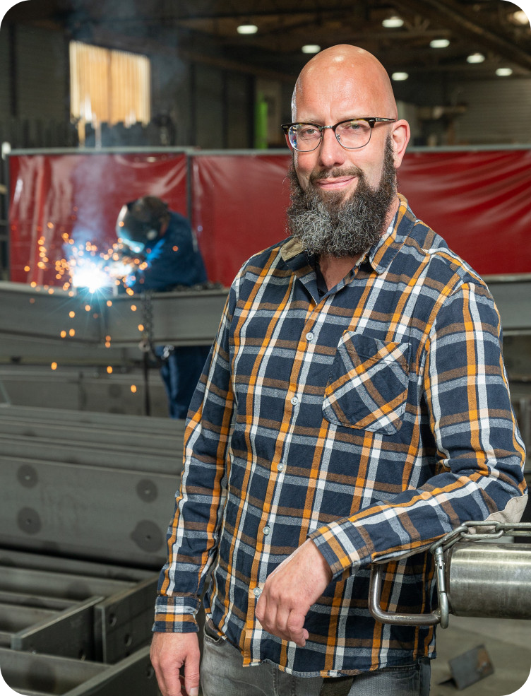 Een man met baard en bril staat voor een lasapparaat in een fabriek. John de Haas