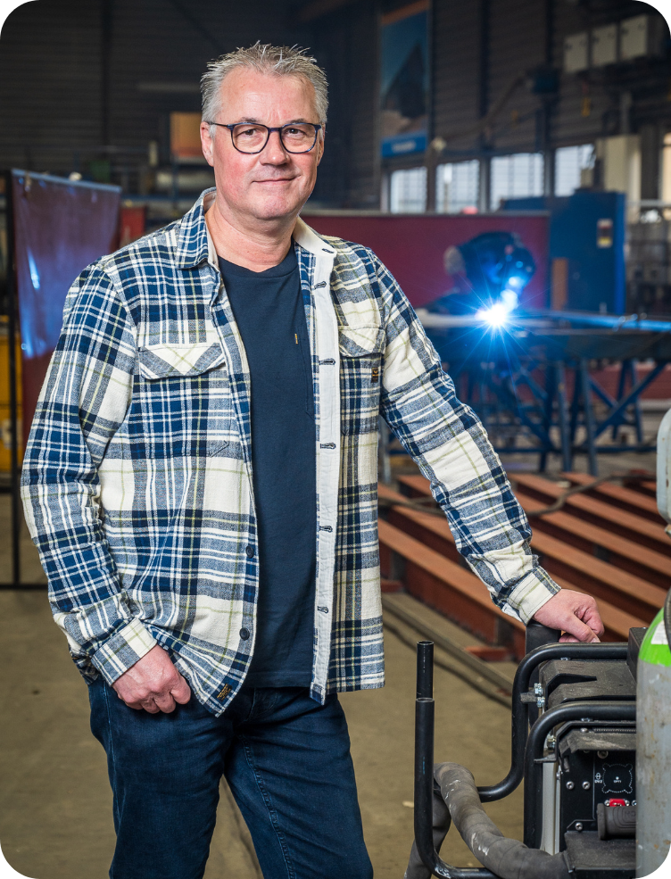 Een jonge man in een groen jasje en spijkerbroek staat in een fabriek. Bas Lamers