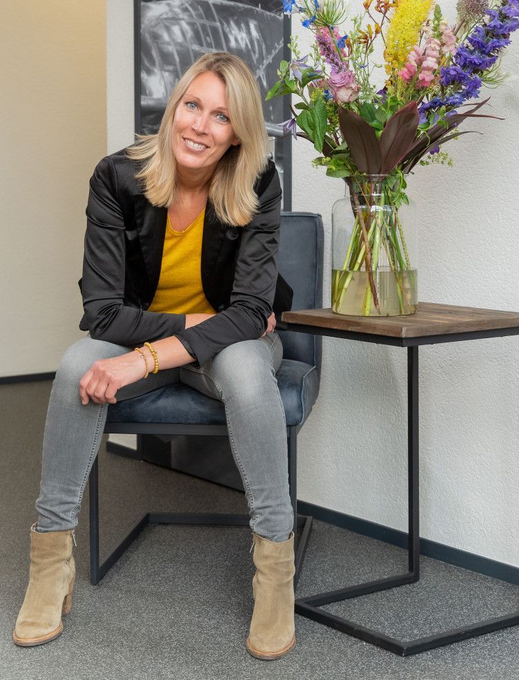 Een vrouw zit op een stoel naast een vaas met bloemen. Anke Vos. 