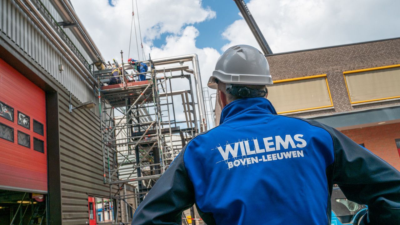 Voor een gebouw in aanbouw staat een man met een harde hoed en een willemsjasje op.