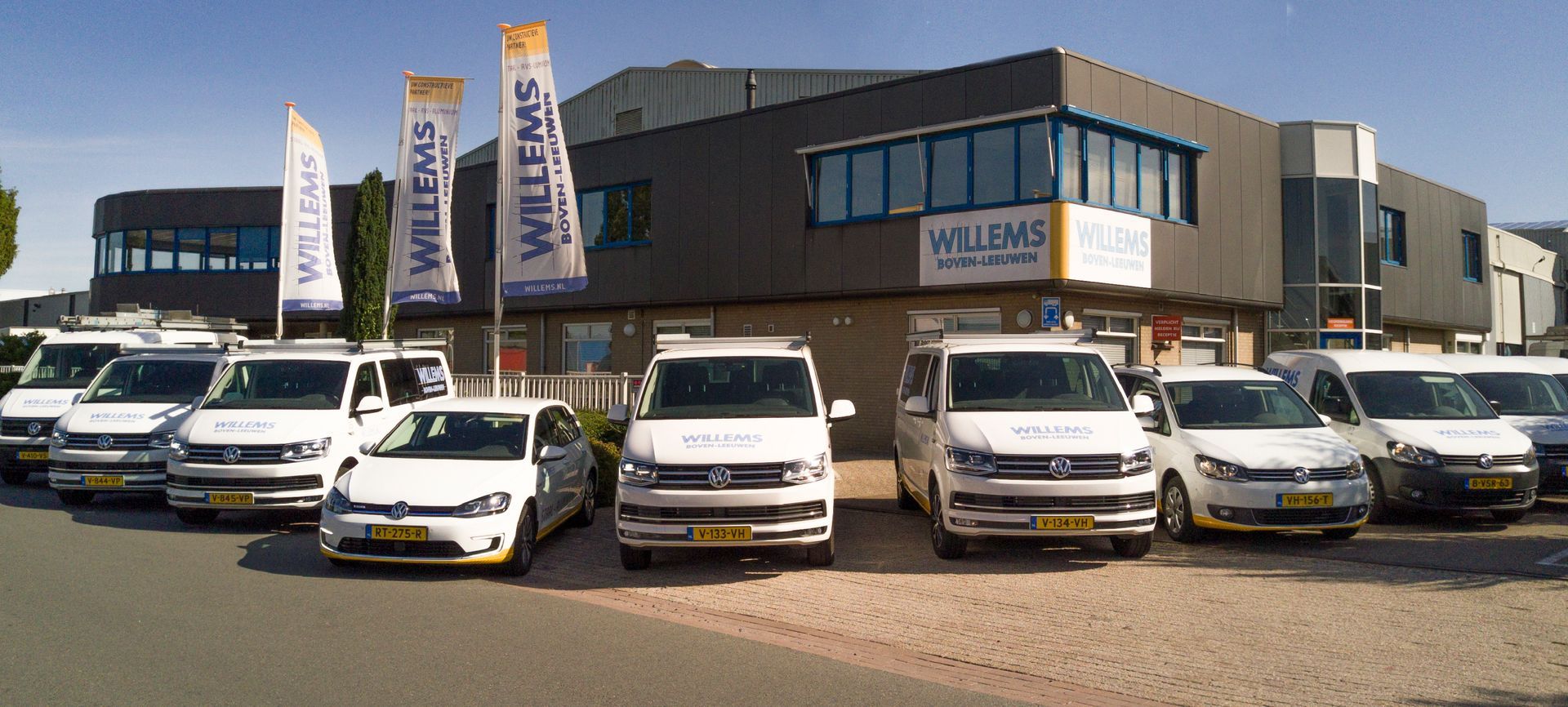 Voor een gebouw met de tekst willems staat een rij witte busjes geparkeerd