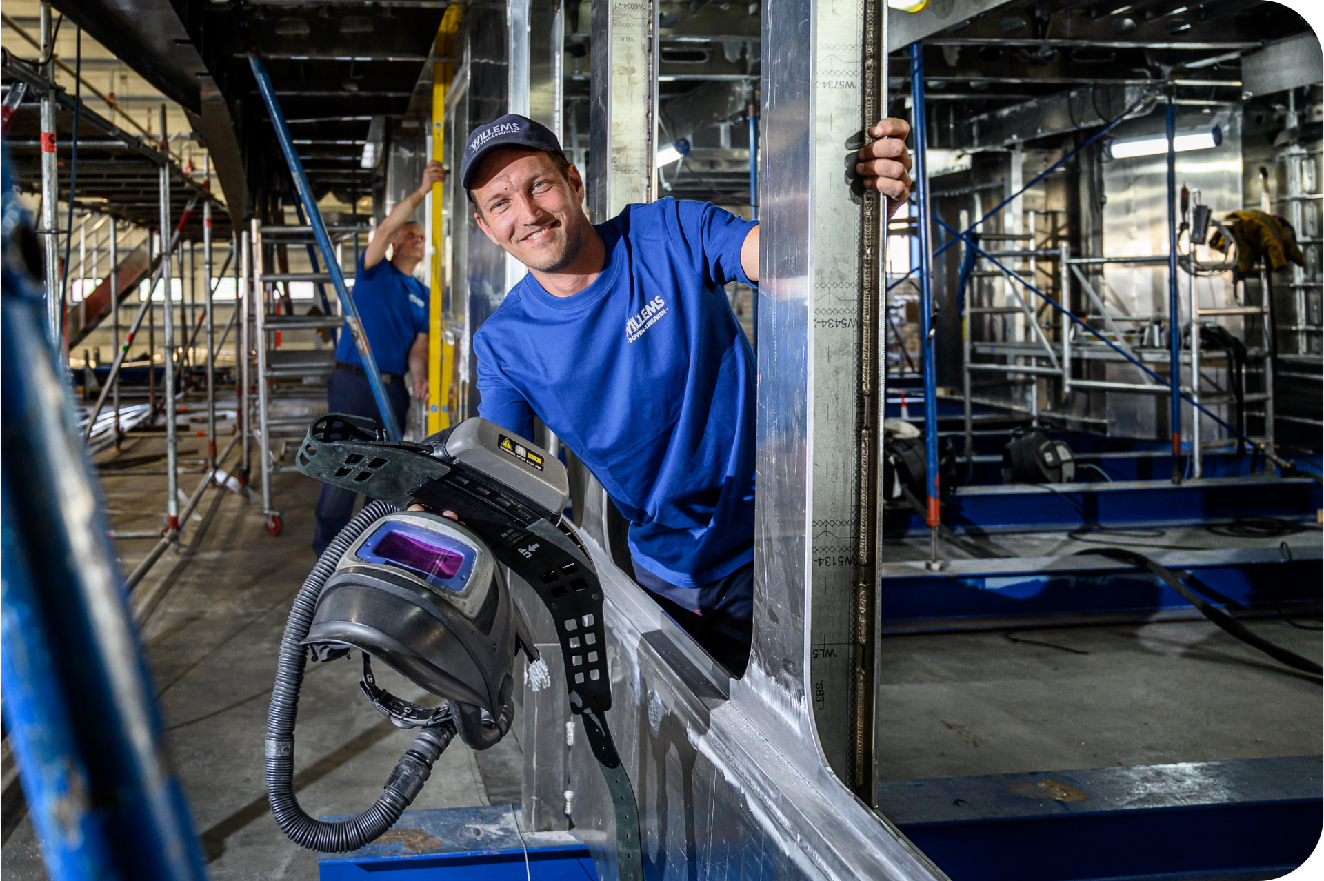 Een man in een blauw shirt staat in een fabriek met een lasmasker in zijn hand. Door Willems Boven-Leeuwen.