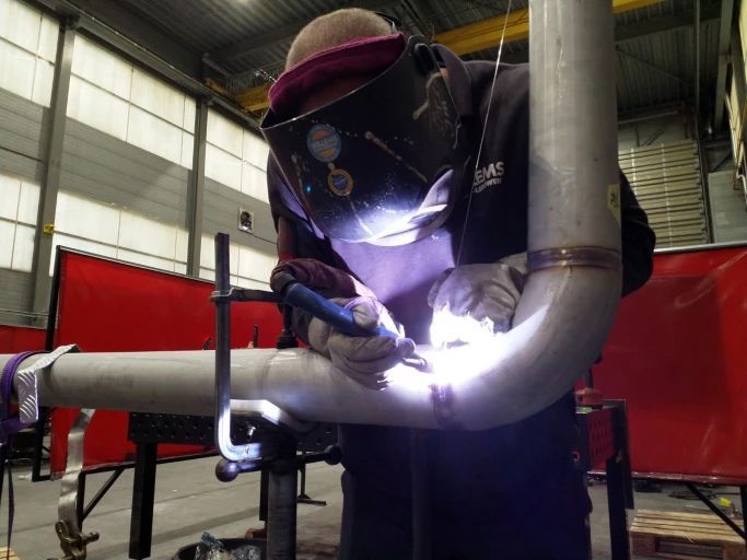 Een man met een lasmasker is een pijp aan het lassen in een fabriek.