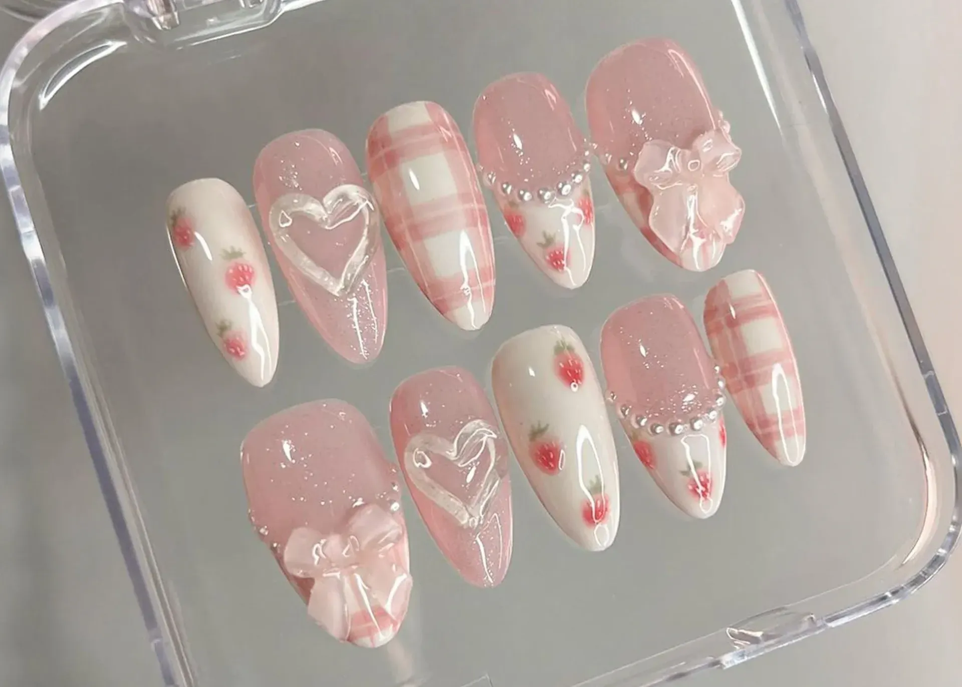 Decoración de uñas rosa y blanca con diseños de fresas, corazones y cuadros, expuesta en un recipiente transparente.