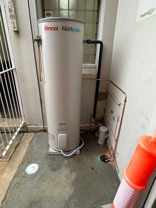 A White Water Heater Is Sitting In A Corner Of A Room Next To A Window — Gordo's Plumbing Service In Wagga Wagga, NSW