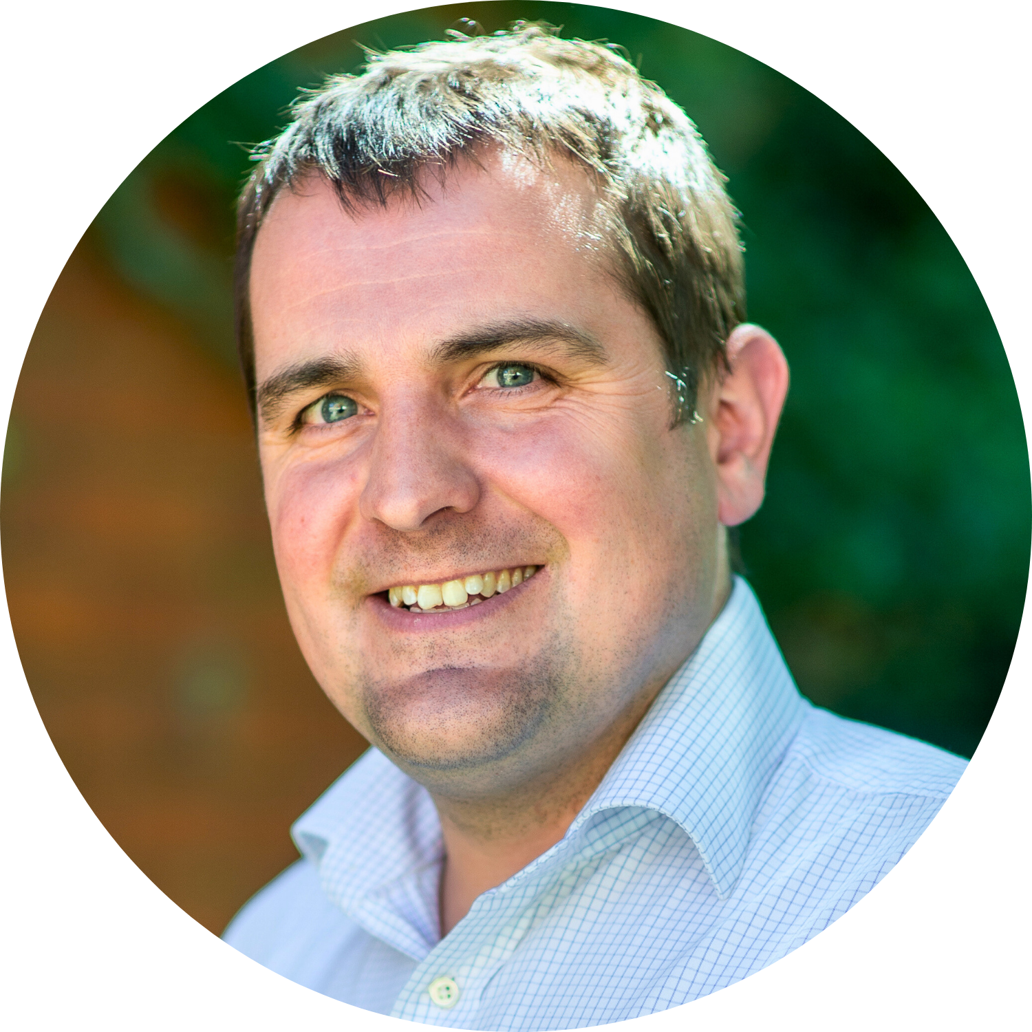 Smiling man in a light blue collared shirt, against a blurred outdoor background.