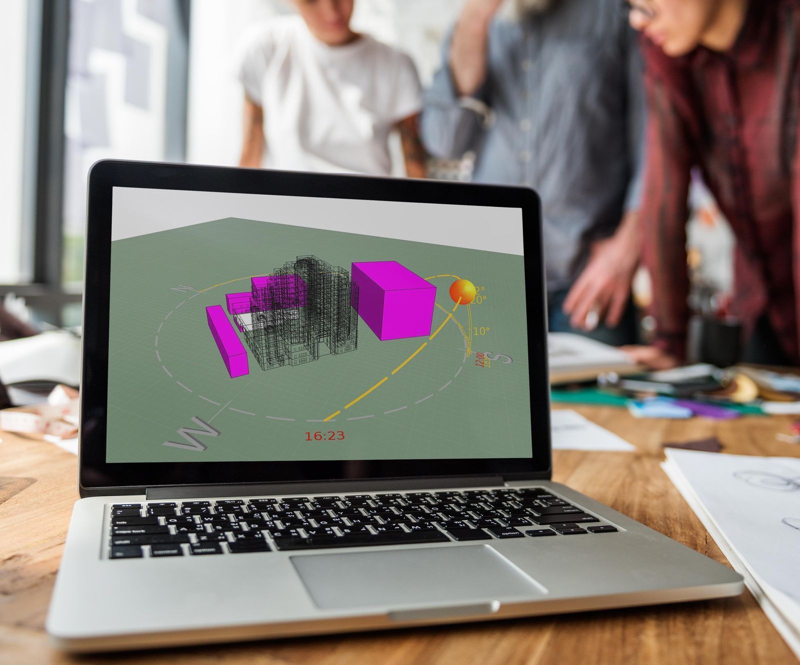 An open laptop displays a digital building model showing the path of the sun around a building. Behind the laptop three people stand around the table, blurred out. 