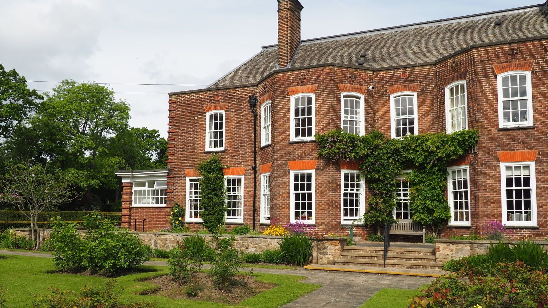 A large Victorian red brick property overlooks a formal garden with large sash windows and a climbing plant. 