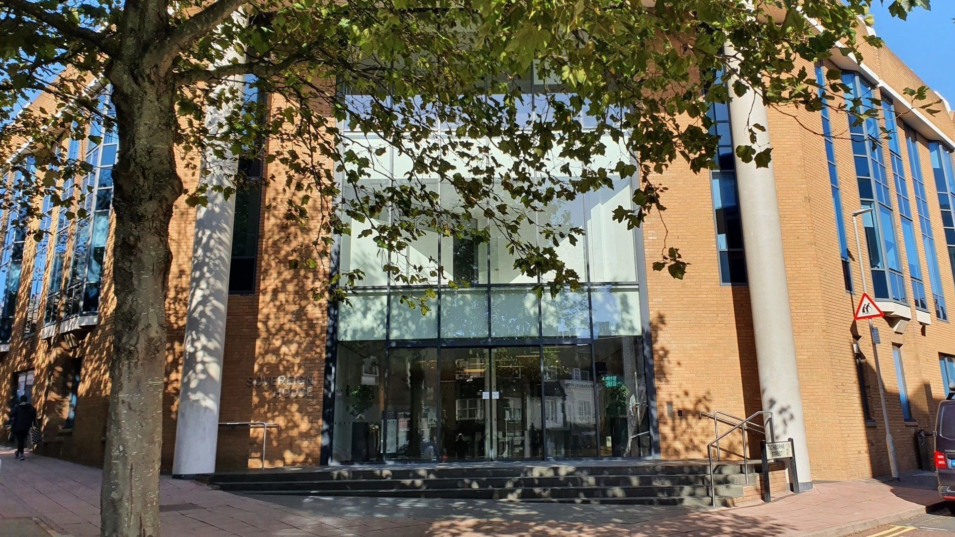 The sun shines on the entrance to a large office building. A tree in leaf sits in the forefront, shading the entrance. 