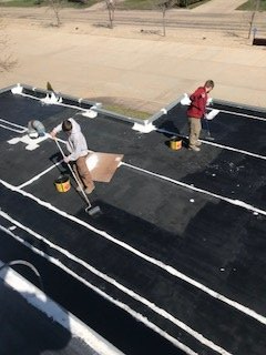 Two workers apply white sealant to the seams and edges of a black flat roof.