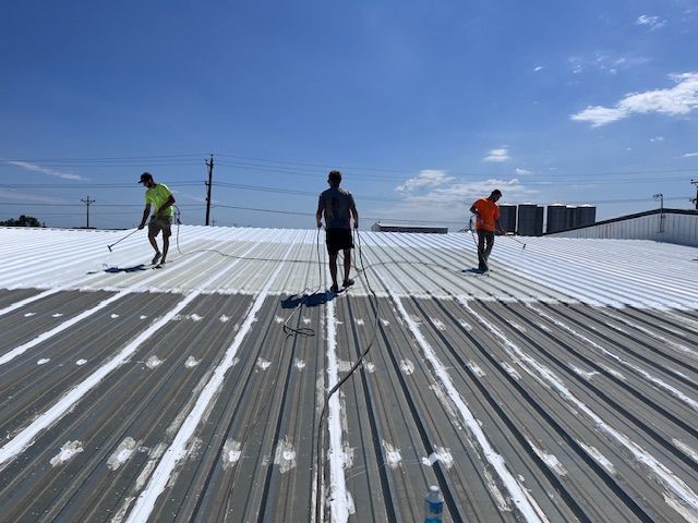 Three workers wearing high-visibility shirts apply a white reflective coating to a corrugated metal industrial roof.