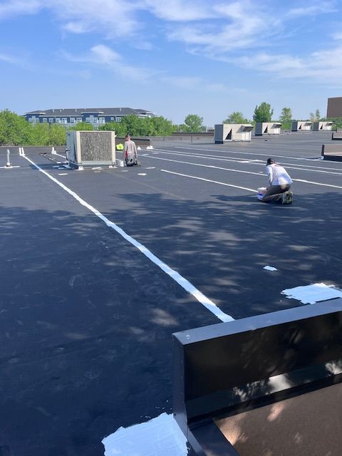Two people working on a large black flat roof with white adhesive lines and HVAC units on a sunny day.