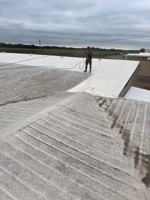 A person using a spray hose to coat a section of a large, gray industrial metal roof with a bright white sealant.