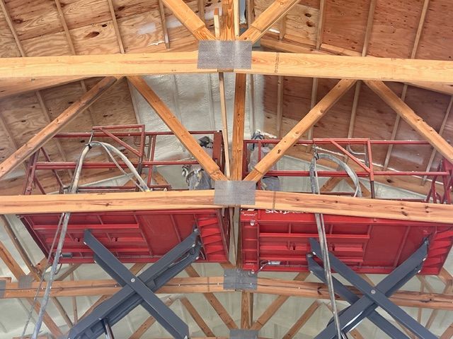 Two red scissor lifts extended upward in a wooden-beamed building with spray-foam insulated roof rafters.