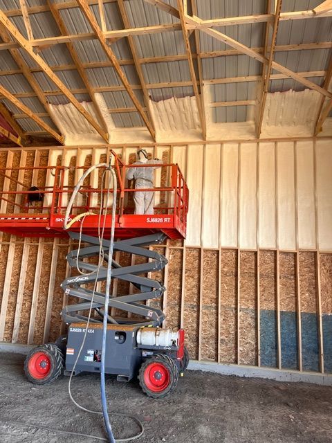 A worker in protective gear on a scissor lift sprays foam insulation onto the wall and ceiling of an unfinished building.