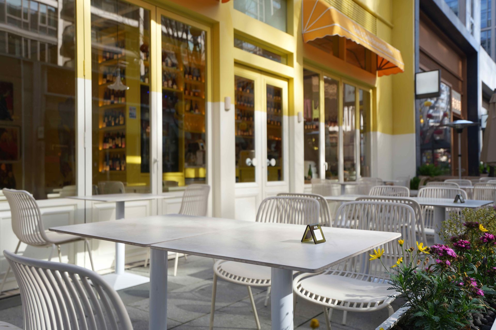 Yellow café exterior with yellow awning, white outdoor tables, chairs, and flower bed. Yellow café exterior with yellow awning, white outdoor tables, chairs, and flower bed.