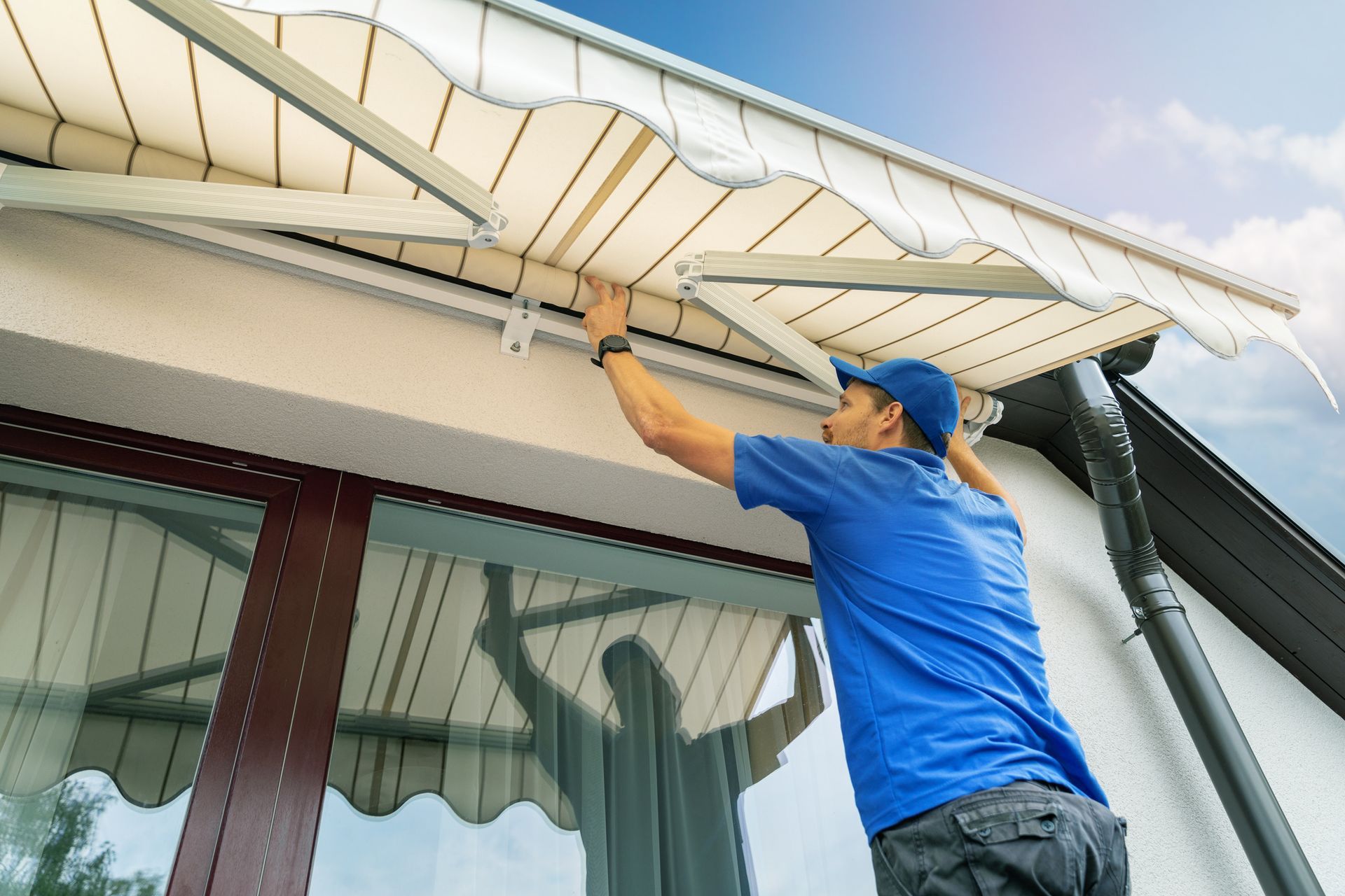 A worker in a blue uniform, installing a residential awning on the house over the terrace window.