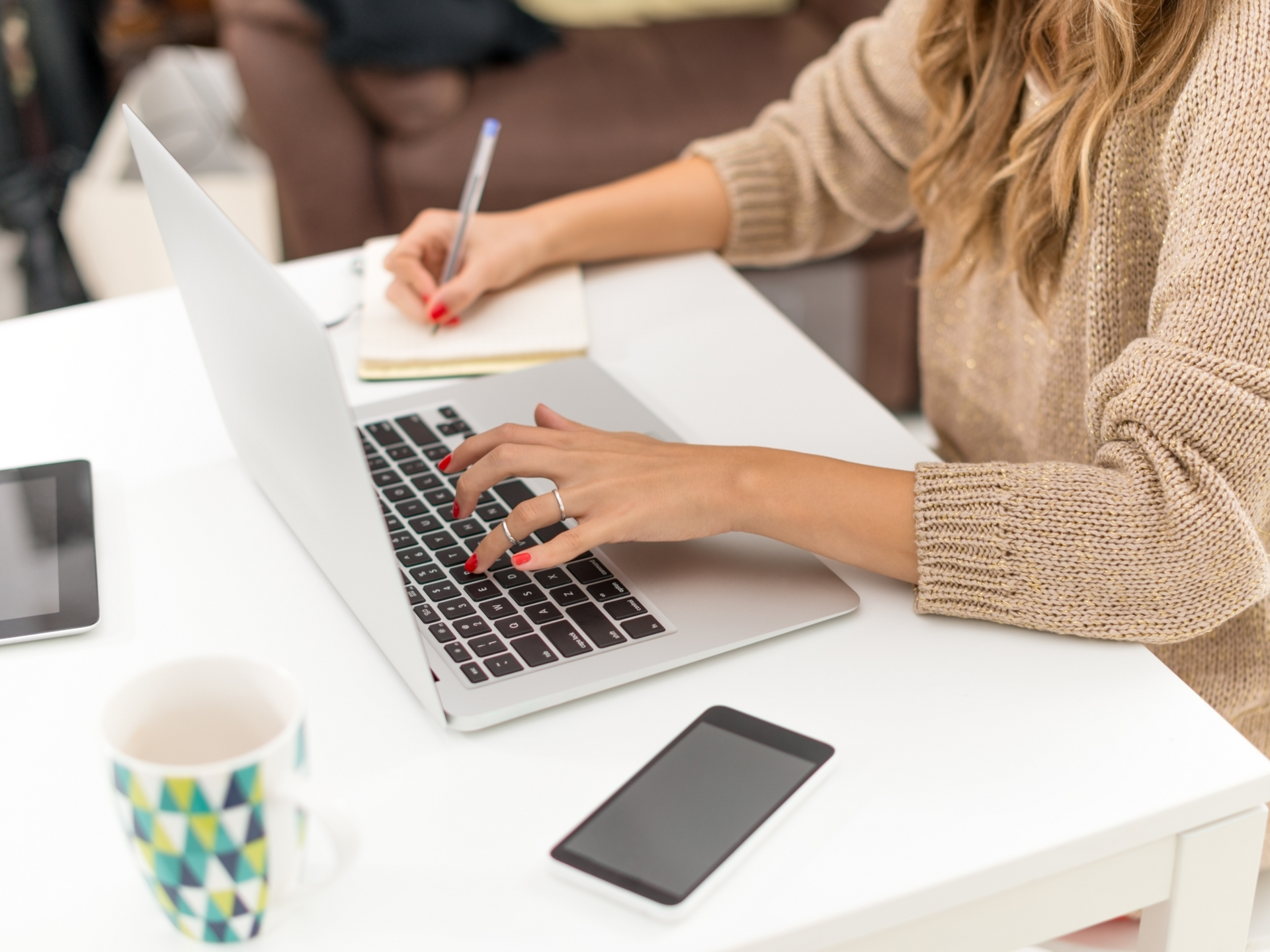A woman is typing on a laptop computer while writing in a notebook.