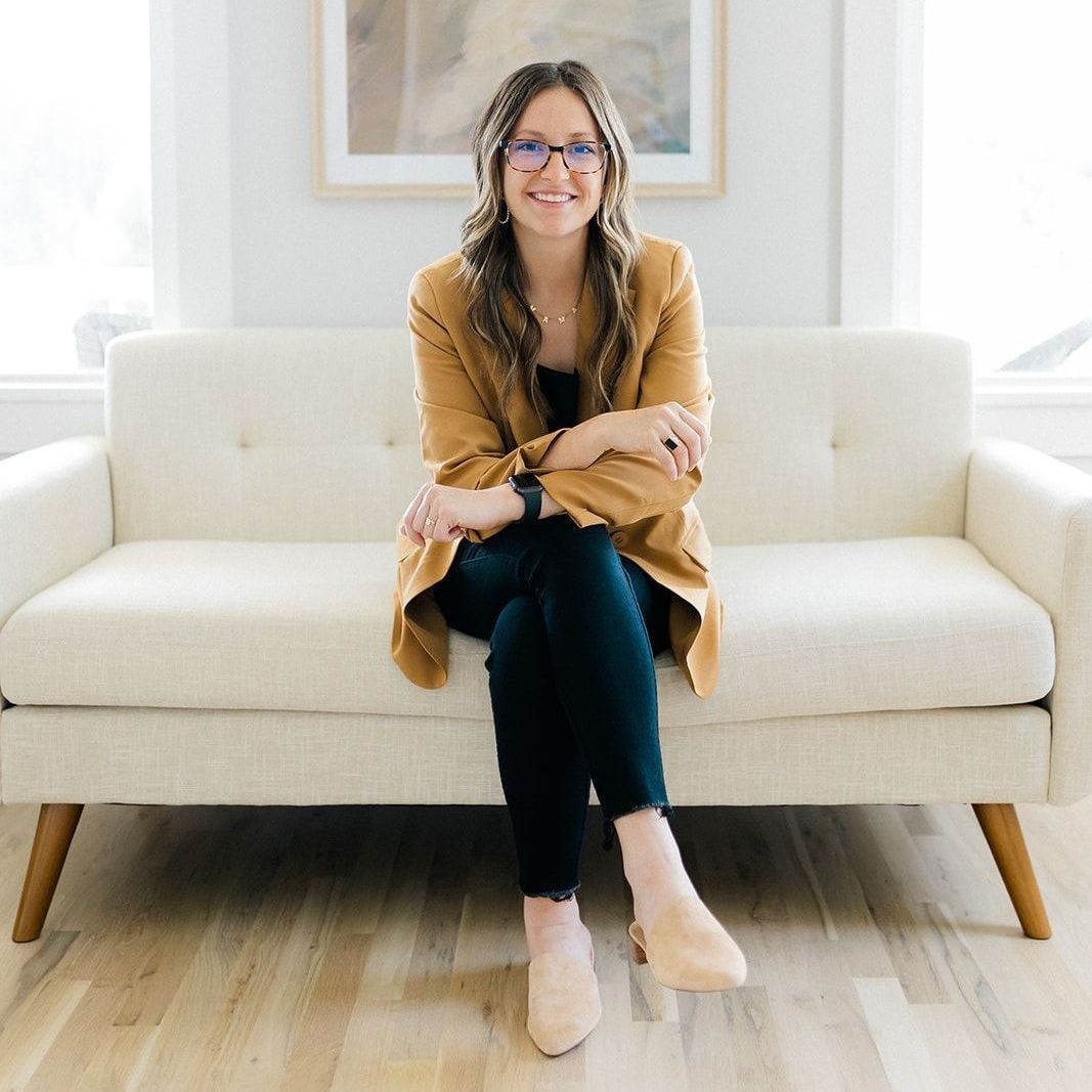 A woman is sitting on a couch wearing glasses and a tan jacket.