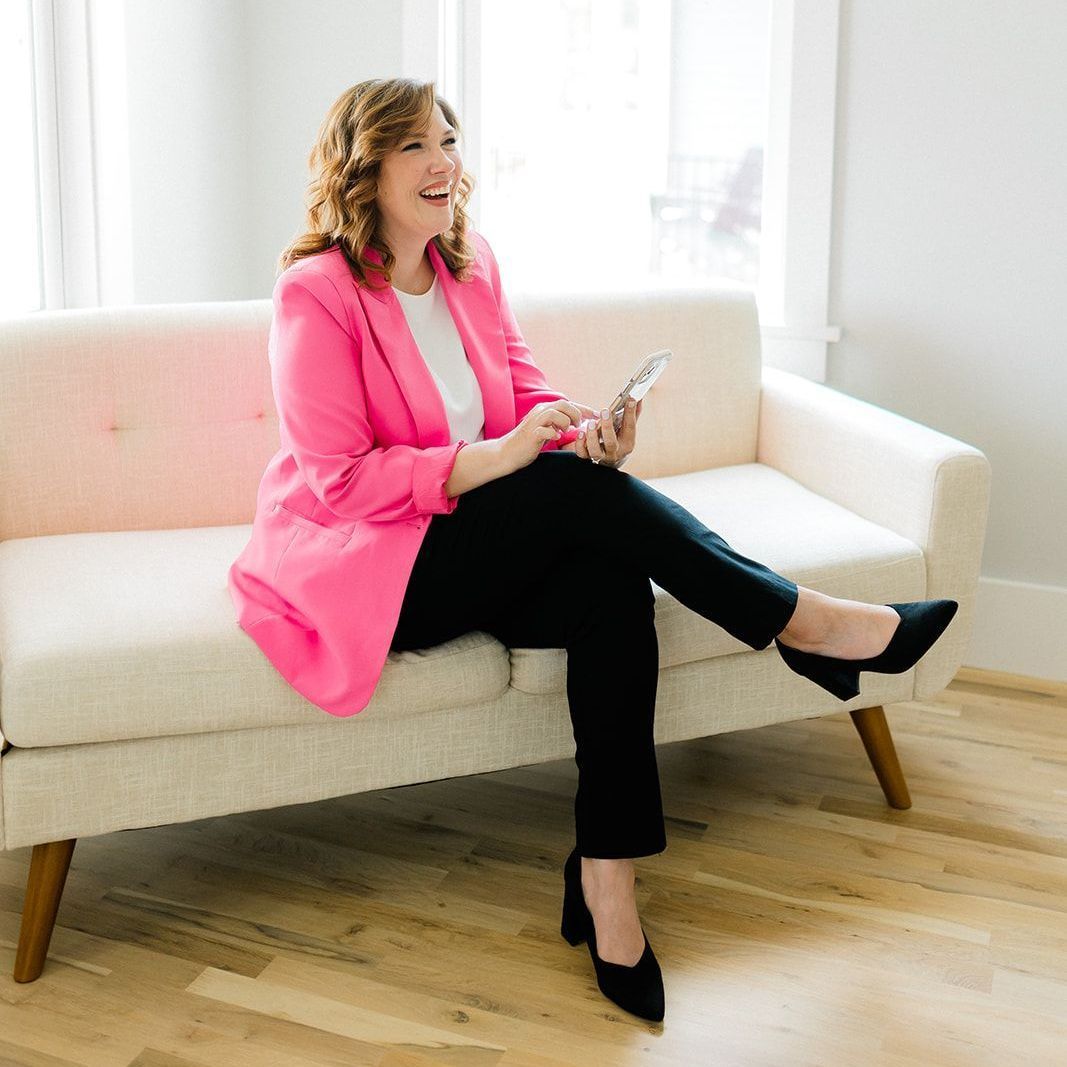 A woman in a pink jacket is sitting on a couch holding a cell phone.