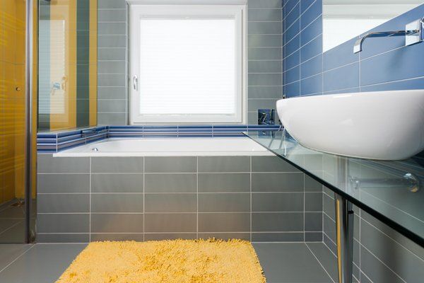 Sink in the bathroom standing on a glass shelf with modern pipe on the wall, bathtub, shower cabin and window in the background