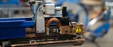 Close-up of an industrial machine component, featuring electrical components, copper coils, and a warning symbol. The setting appears to be a factory.