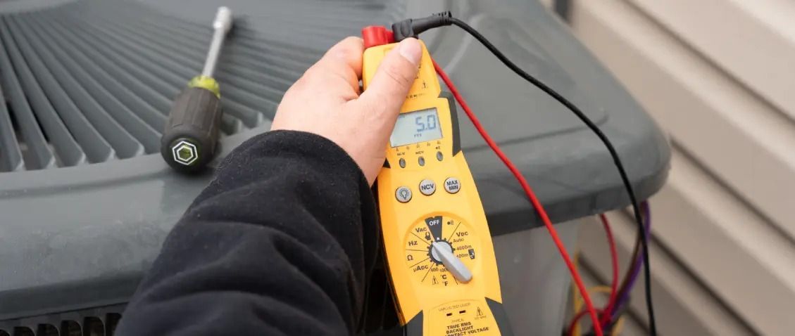 A person uses a yellow multimeter to test an air conditioning unit. A screwdriver sits nearby on the unit's exterior.