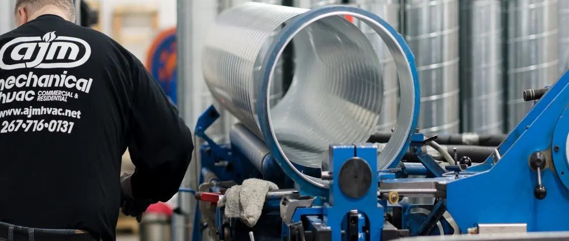 A worker in a black shirt with a company logo works on industrial machinery in a workshop. A large cylindrical metal object is visible.
