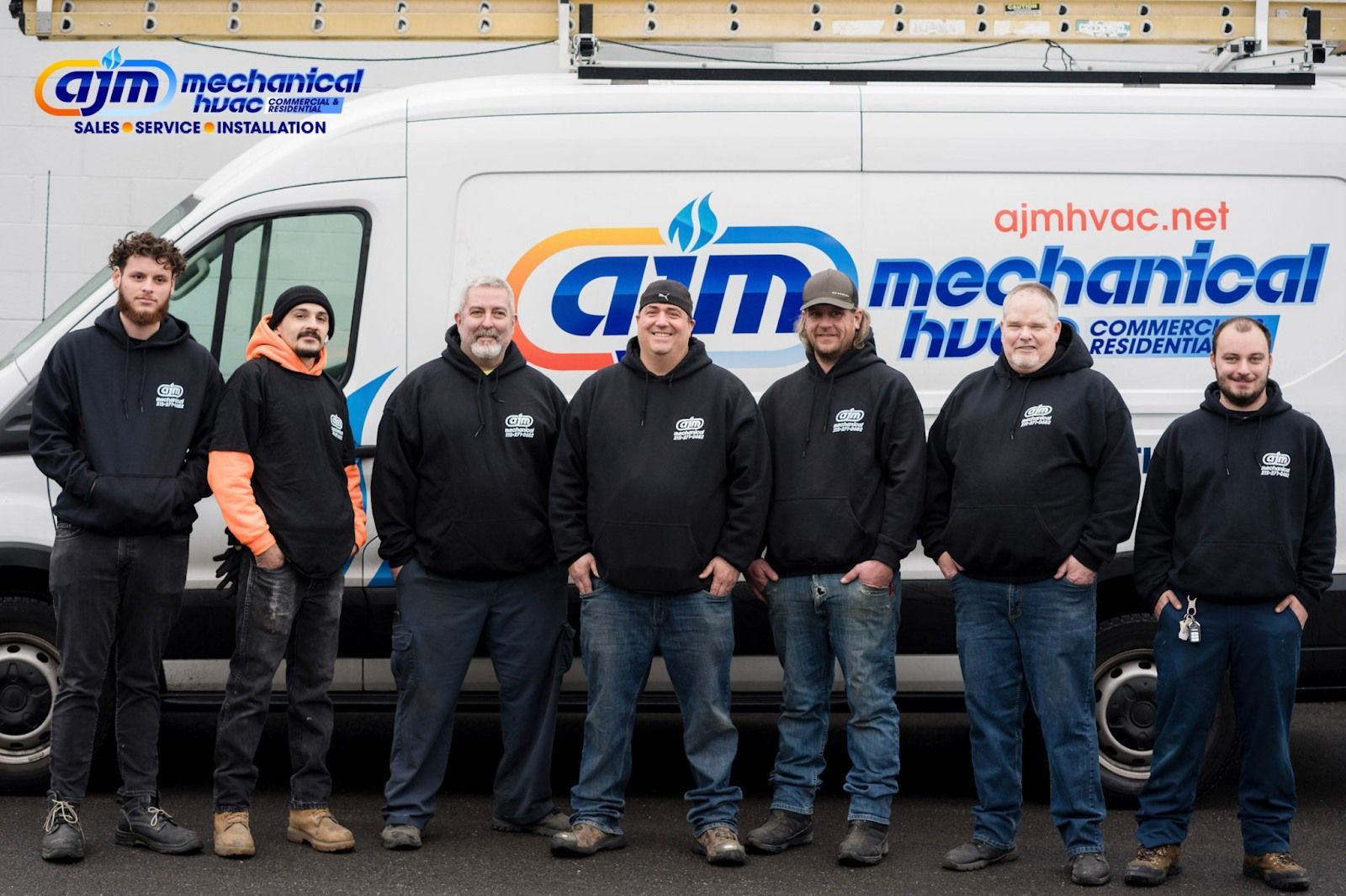 A group of seven men in black hoodies standing in front of a white AJM Mechanical van. They appear to be a work crew.