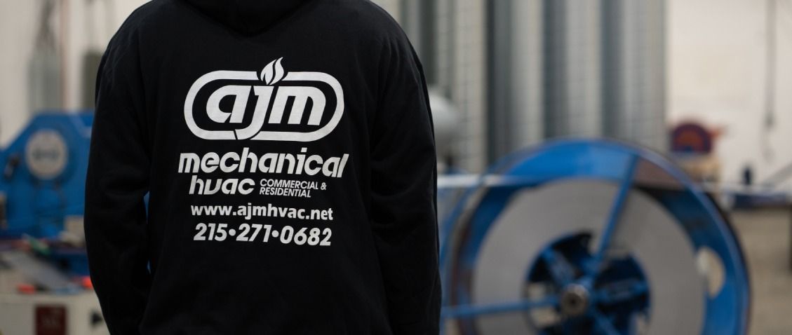 A person wearing a black hoodie with a white AJM Mechanical logo, standing in a workshop with industrial equipment.