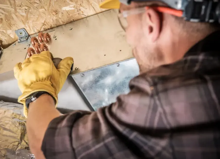 A person in safety glasses and gloves installs ductwork in an unfinished attic, near wooden beams.