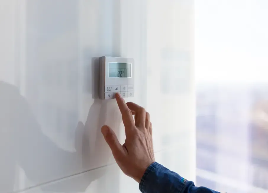A person's hand adjusting a digital thermostat on a white wall near a window.