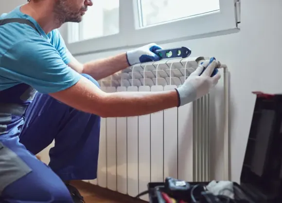 A person wearing work gloves kneels to check a radiator with a level near a window. They wear a blue shirt and overalls.