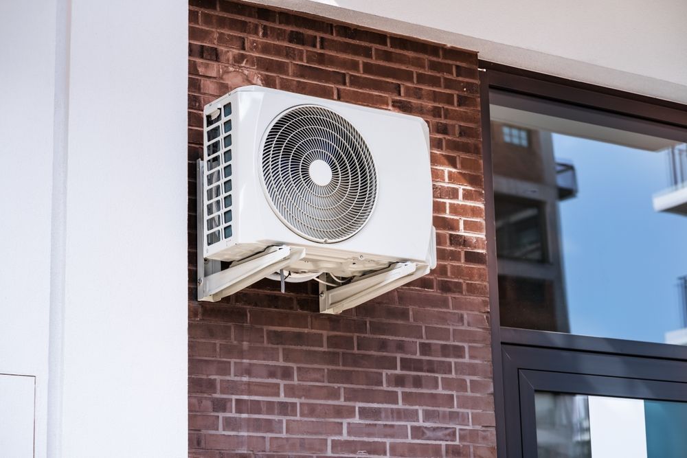 An outdoor air conditioning unit, mounted on a red brick wall next to a window. The unit is white.
