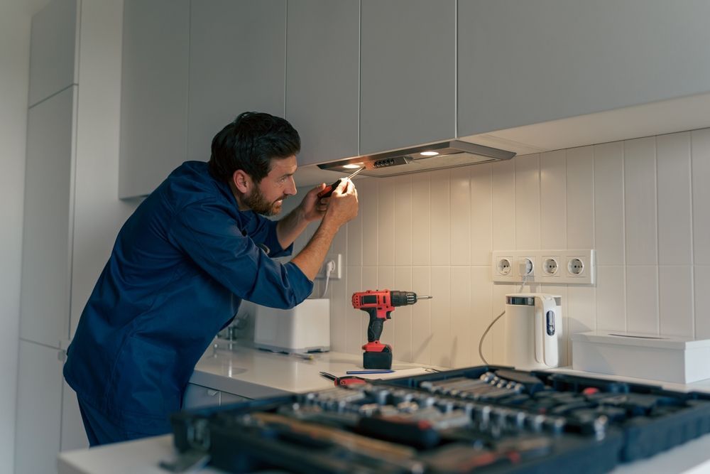 A man in a blue uniform repairs a range hood in a modern kitchen. He is focused, holding a small tool near the light.
